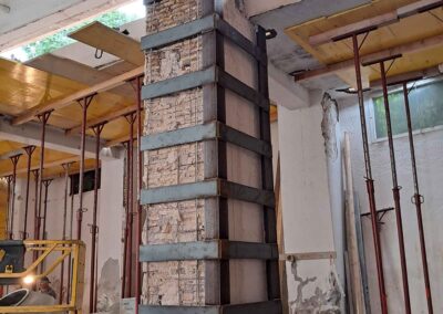 Una colonna di cemento in un edificio in fase di ristrutturazione è rinforzata con fasce di acciaio e supporti temporanei. L'area circostante, gestita da stepa.it, mostra travi a vista, impalcature e materiali da costruzione.